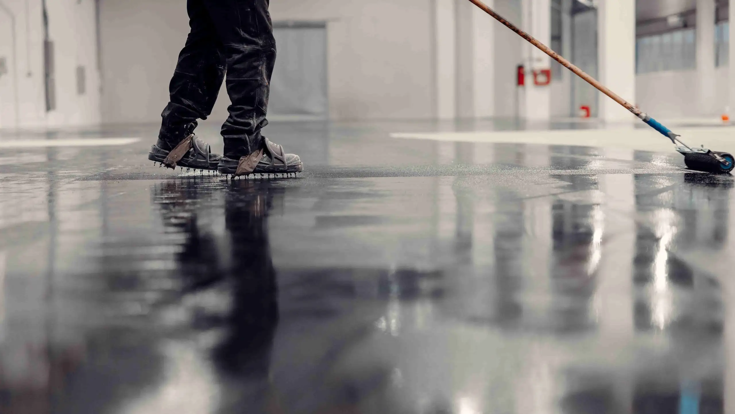 Worker applying epoxy coating on the industrial floor with a roller, reflecting surface, in a modern warehouse setting.